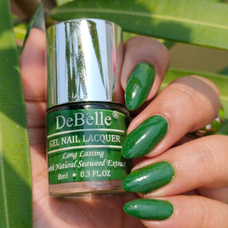 Holding DeBelle Dark jade Green nail polish with a beautifully manicured nails against a green leaves background.