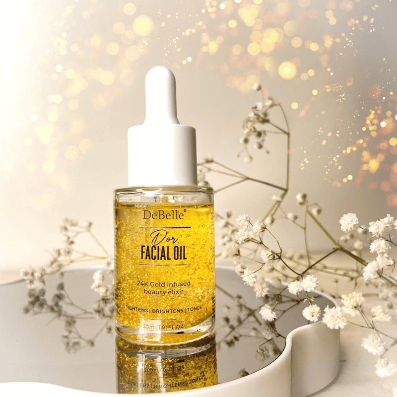Debelle Facial Gold Oil: Luxurious bottle of gold-infused facial oil surrounded by flowers on a pristine glittery background shiny surface