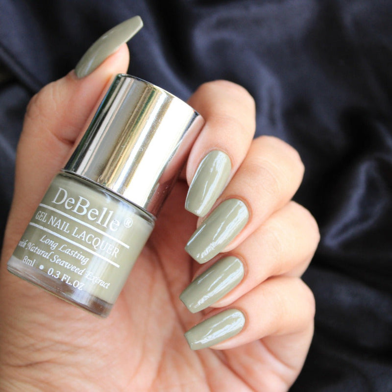 Holding DeBelle Olive Green nail polish with a beautiful manicured nails against a dark blue background.