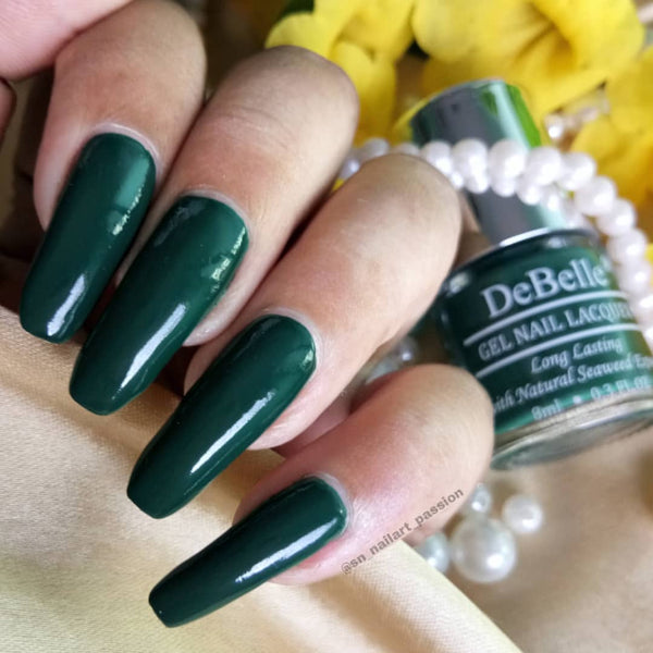 Close-in view of a nail painted with DeBelle Dark green nail polish against a yellow background.