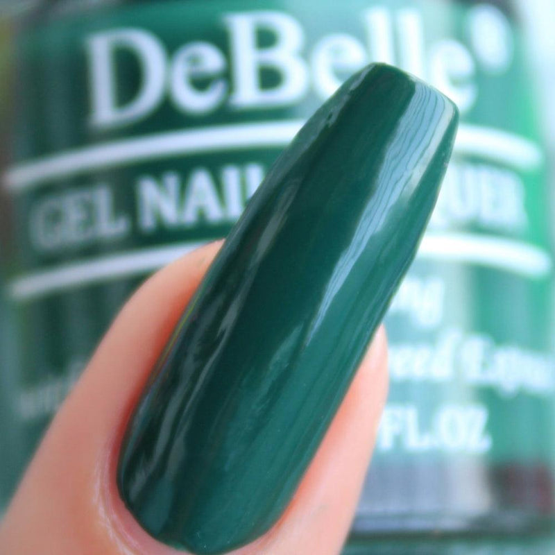 Close-in view of a nail painted with DeBelle olive green nail polish against a blurry green background..