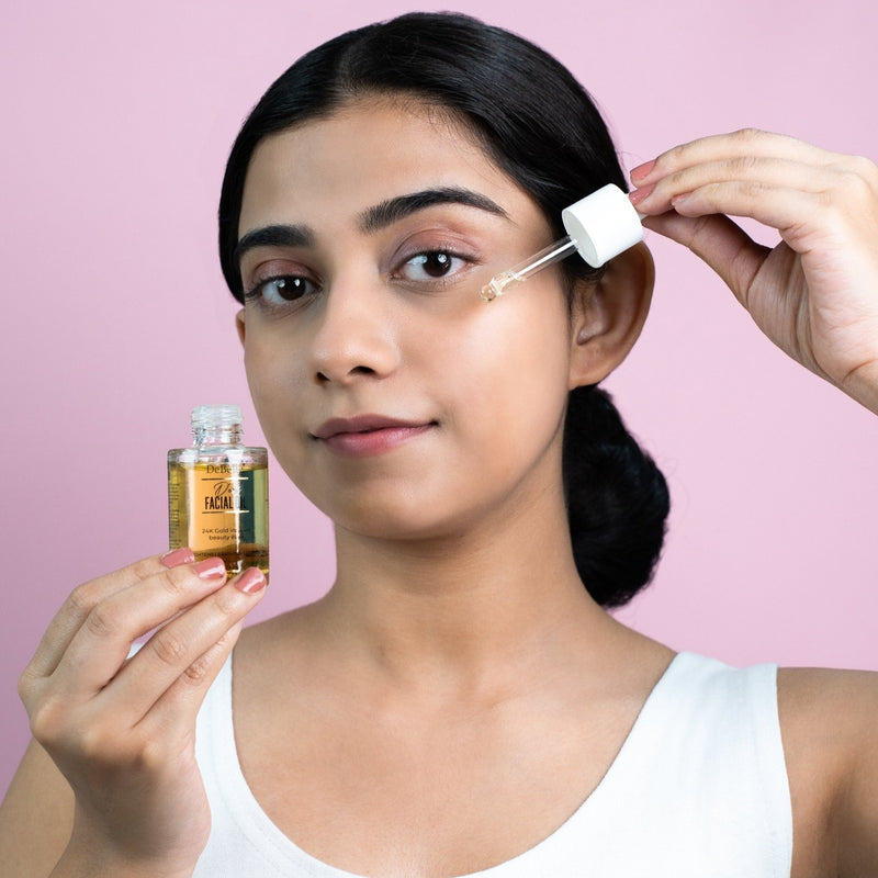 Girl applying Debelle Gold facial oil holding the bottle against a pink background.