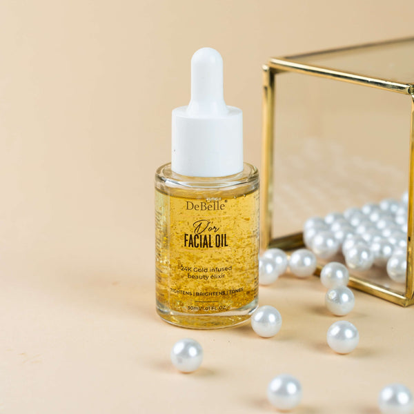 DeBelle Facial Gold Oil: Golden bottle of nourishing facial oil placed on a cream surface against a backdrop shinny pearl.