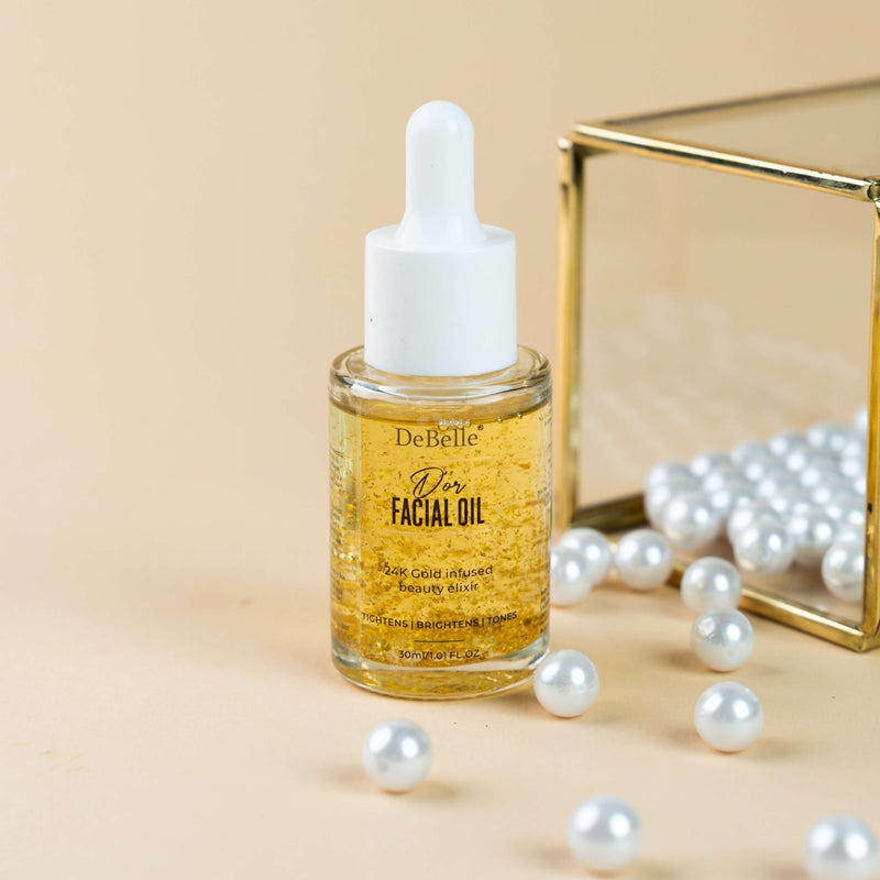 DeBelle Facial Gold Oil: Golden bottle of nourishing facial oil placed on a cream surface against a backdrop shinny pearl.