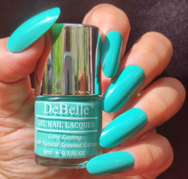 Holding DeBelle Turquoise jade Green nail polish with a beautifully manicured nails against a maroon background.