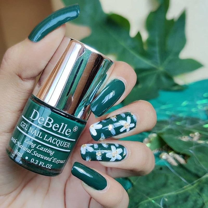 Holding DeBelle Dark Green nail polish with a beautifully manicured nails against a green Leafs background.