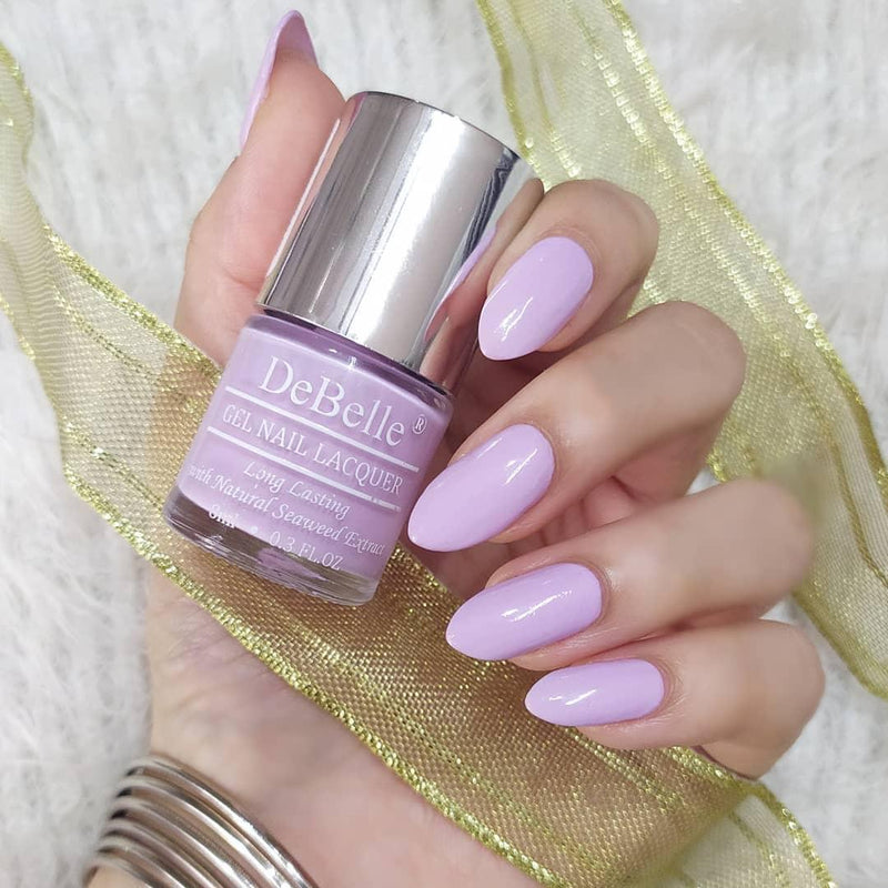 DeBelle Soft lilac lavender Nail Polish - Close-up view of the nail polish bottle and manicured nails against a cream background with a golden ribbon 