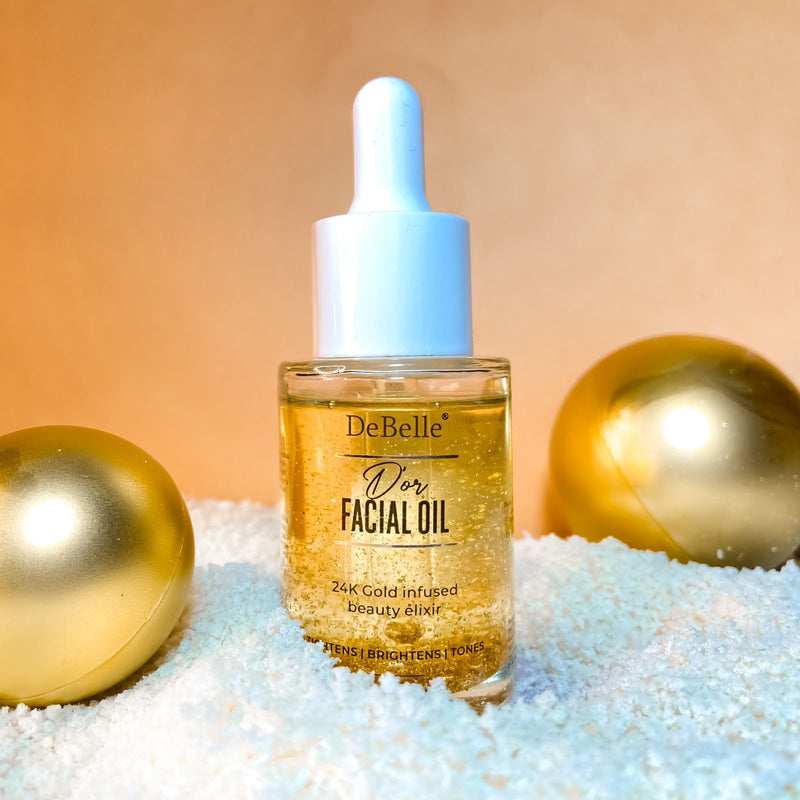 Front view of Debelle gold facial oil with two golden balls and a white surface against a light orange background.