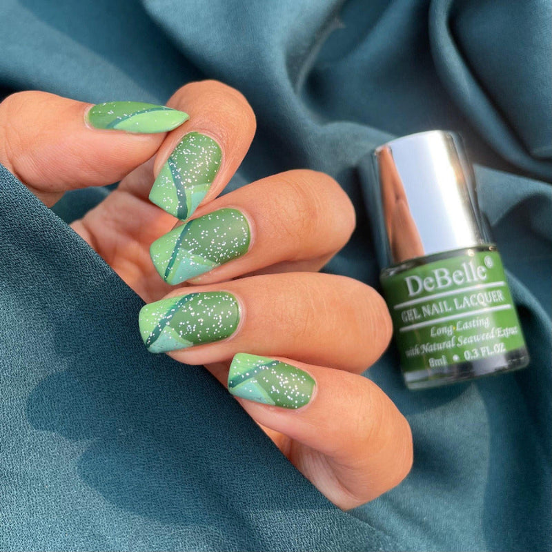 Close-in view of a nail painted with DeBelle dark jade green nail polish against a blue background.