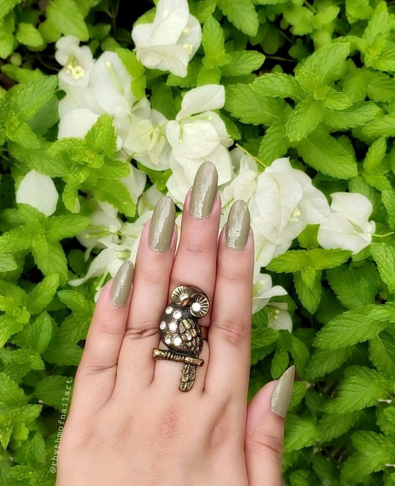 Close-in view of beautifully manicured nails with DeBelle olive green nail polish with green plants in the background.