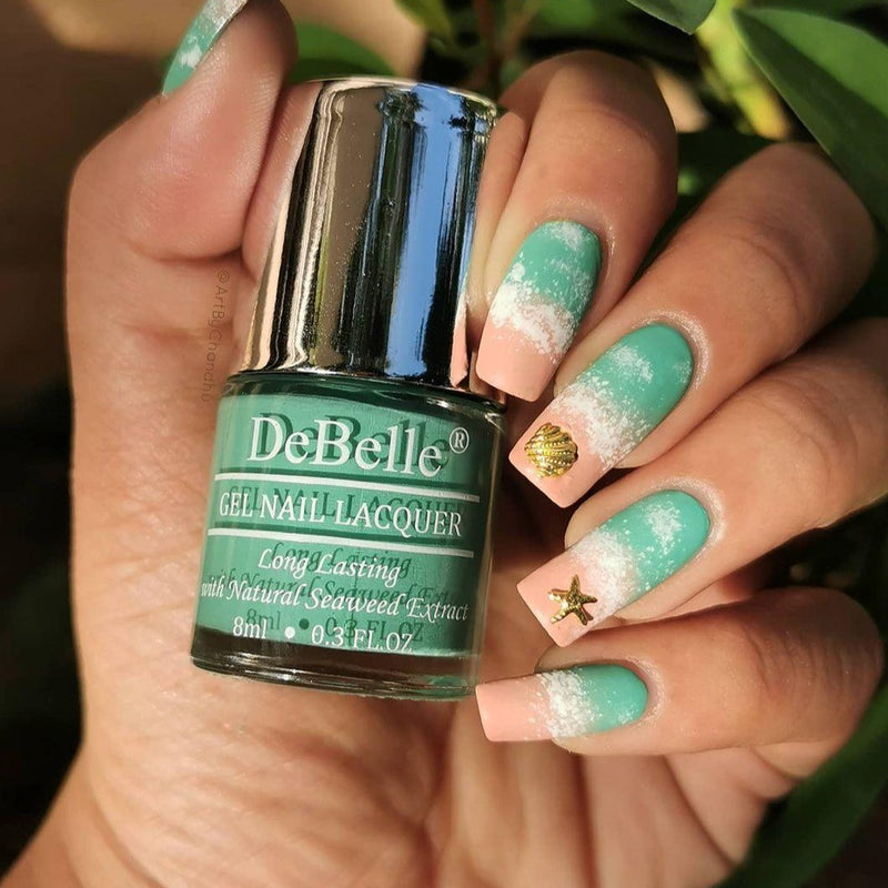 Holding DeBelle Turquoise jade Green nail polish with a beautifully manicured nails against a green leaves background.