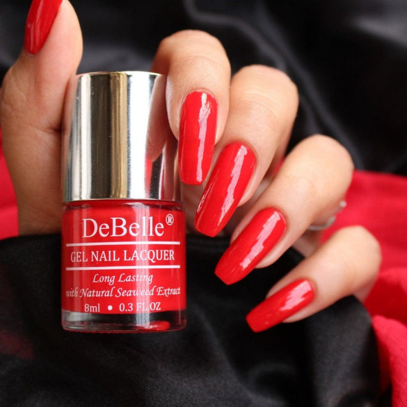 Holding a Scarlet Red Nail polish Bottle from Debelle with the painted nail of same shade has Dark Red ribbon as a background with dark shade