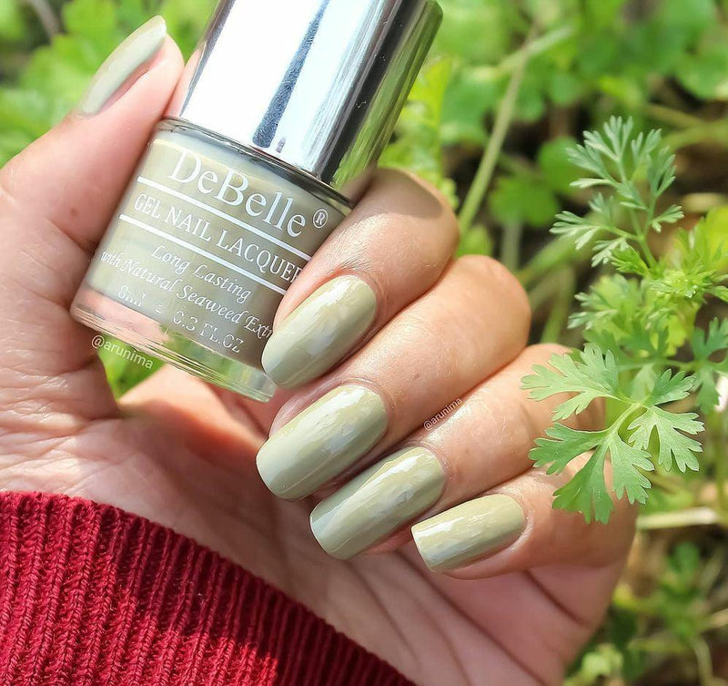 Holding DeBelle Olive Green nail polish with a beautiful manicured nails against a Green plants  background.