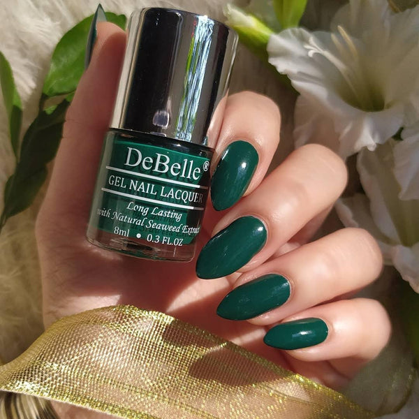 Holding DeBelle Dark Green nail polish with a beautifully manicured nails against a white flowers background.