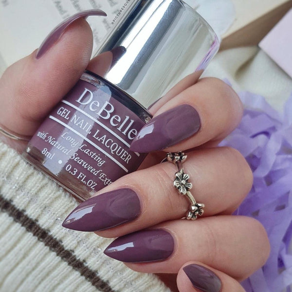 DeBelle Vintage Mauve - Close-up view of the nail polish bottle and manicured nails has purple shaded background 
