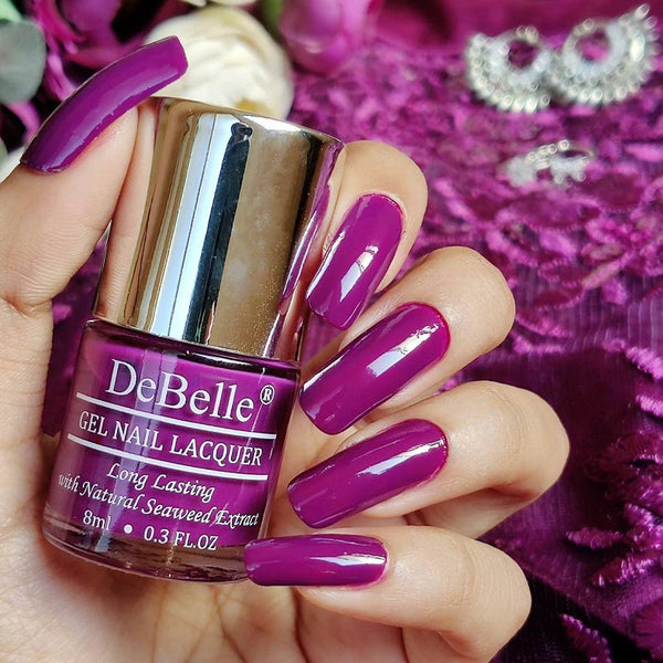 Holding Deep Magenta Nail polish Bottle from Debelle with the painted nail of same shade against a purple background.