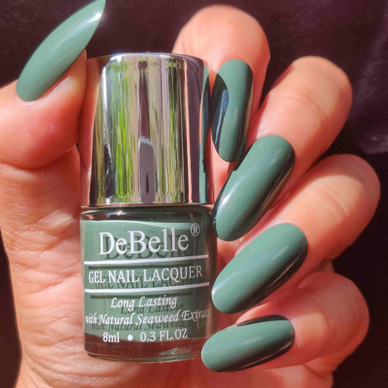 DeBelle green -close-up view of nail polish bottle with the manicured nail with the elegance against a maroon background.