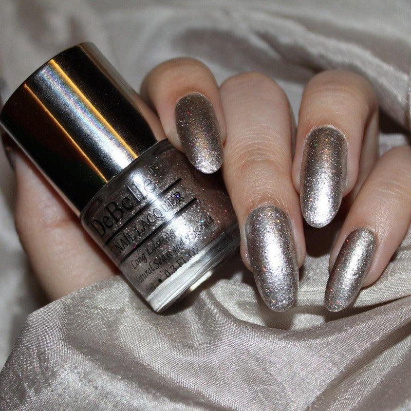 holding a Debelle sparkling dust nail polish with a shiny manicured nails and a silver cloth in the background.