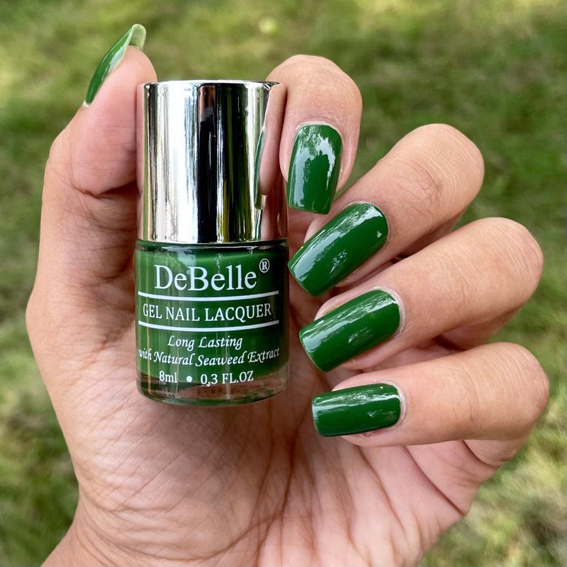 Holding DeBelle Dark jade Green nail polish with a beautifully manicured nails against a green shade background.