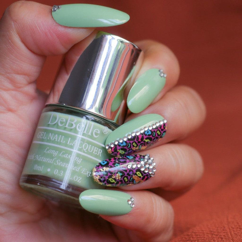 Holding DeBelle mint Green nail polish with a beautifully manicured nails against a red background.